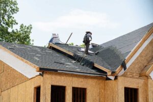 Roofing workers inspect a residential roof deck beside stacked materials and protective ground coverings.