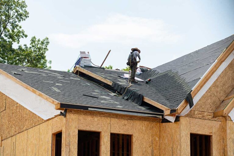 Roofing workers inspect a residential roof deck beside stacked materials and protective ground coverings.